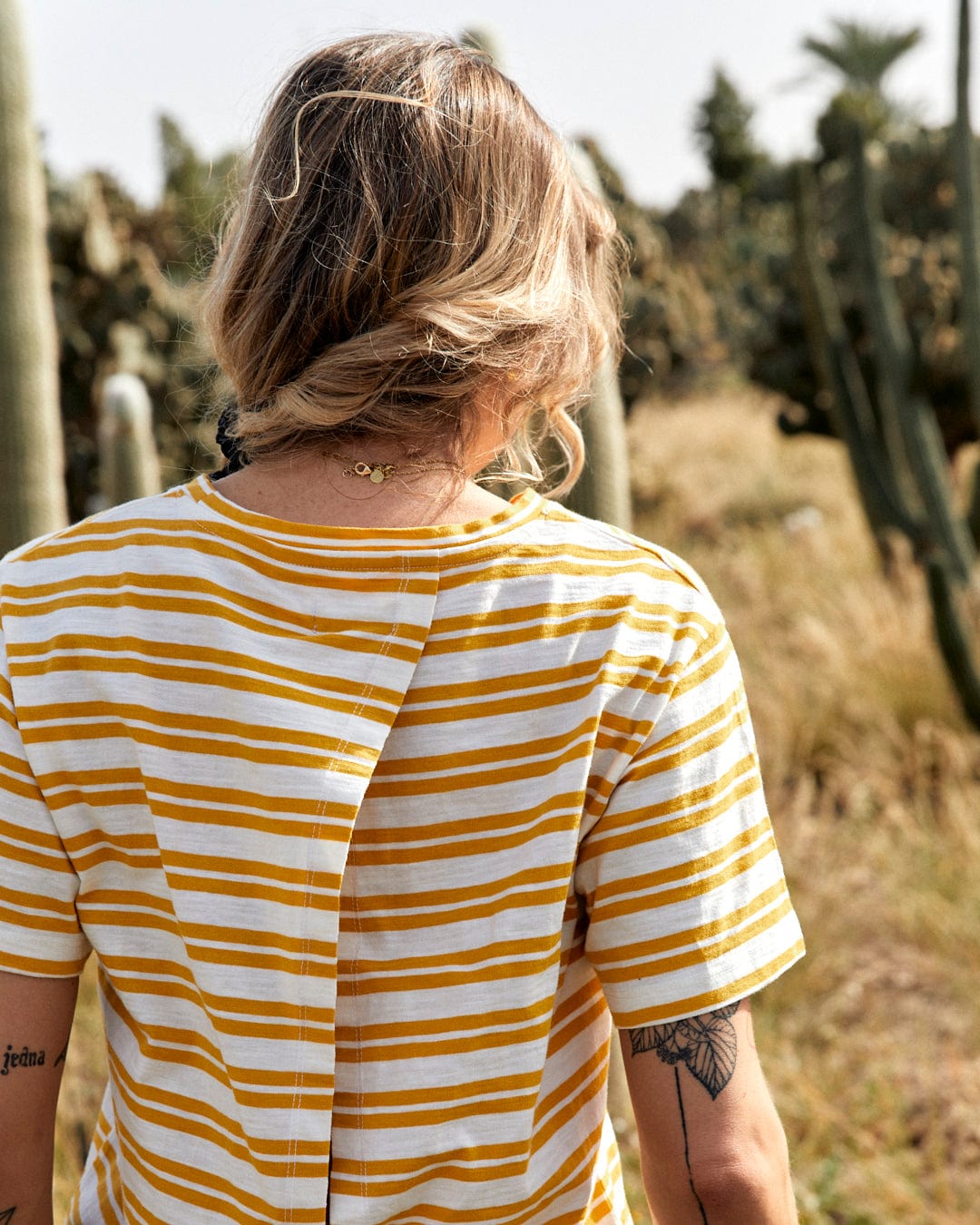 Person with short hair wearing a Saltrock Sky - Womens Short Sleeve T-Shirt in Yellow and white yarn dye stripe print, viewed from behind, walking in a field with tall cacti.