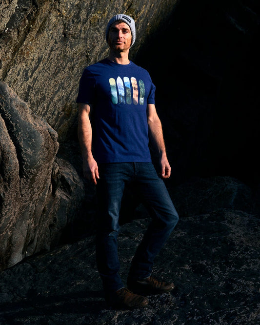 A man wearing a blue t-shirt standing in front of rocks, showcasing Snowboards - Mens Short Sleeve T-Shirt - Blue designs made by Saltrock.