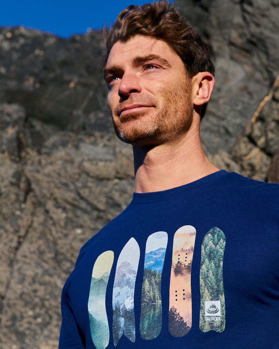 A man wearing a Saltrock Snowboards - Mens Short Sleeve T-Shirt - Blue standing in front of rocks.