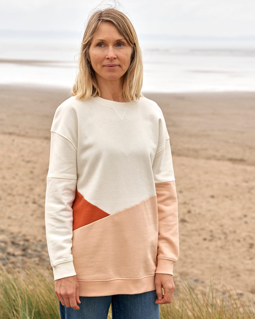A woman standing on a beach wearing the Sofie - Womens Boyfriend Fit Sweat - Light Orange/Cream sweater by Saltrock.