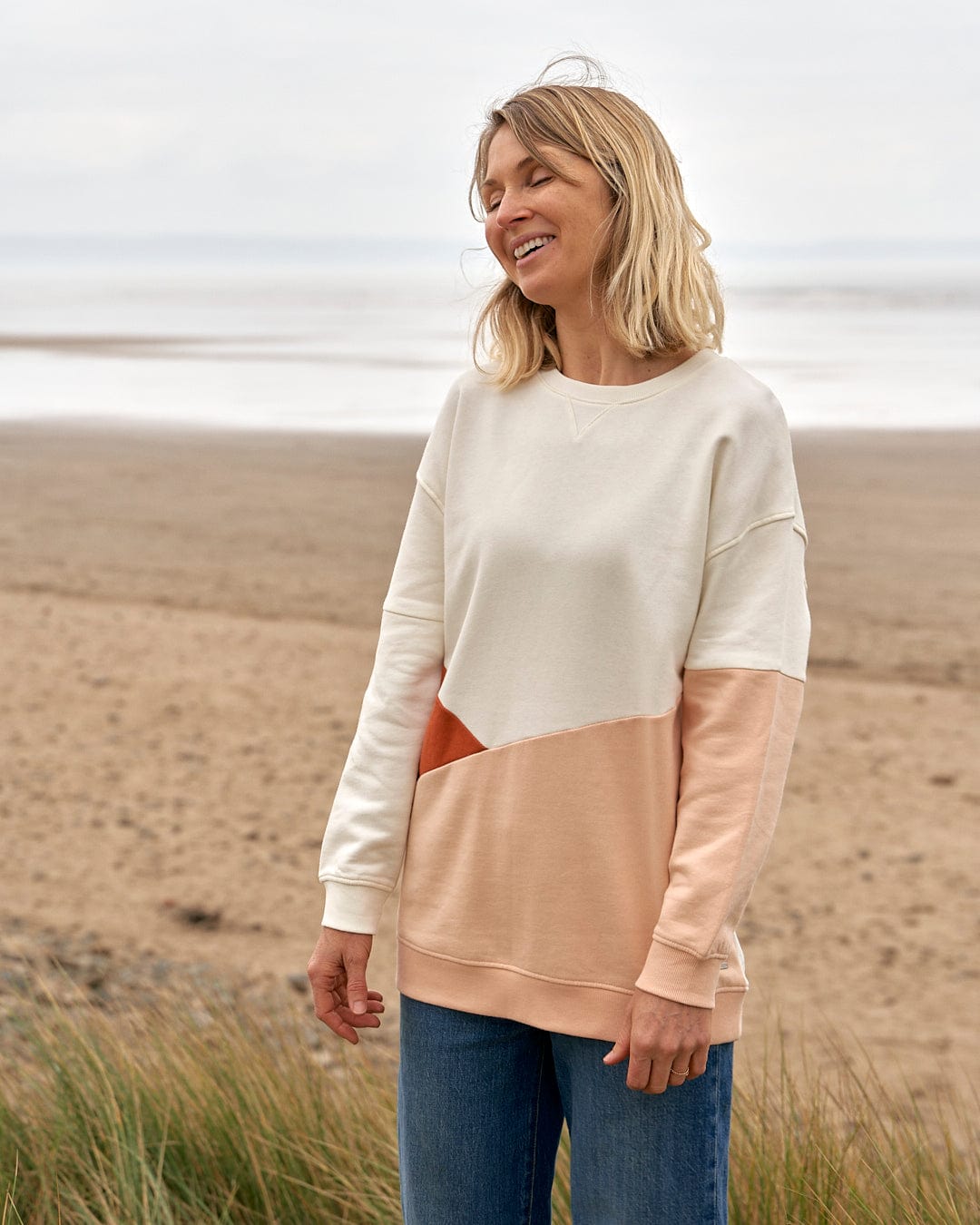A woman standing on a beach wearing a Saltrock Sofie - Womens Boyfriend Fit Sweat - Light Orange/Cream sweater.