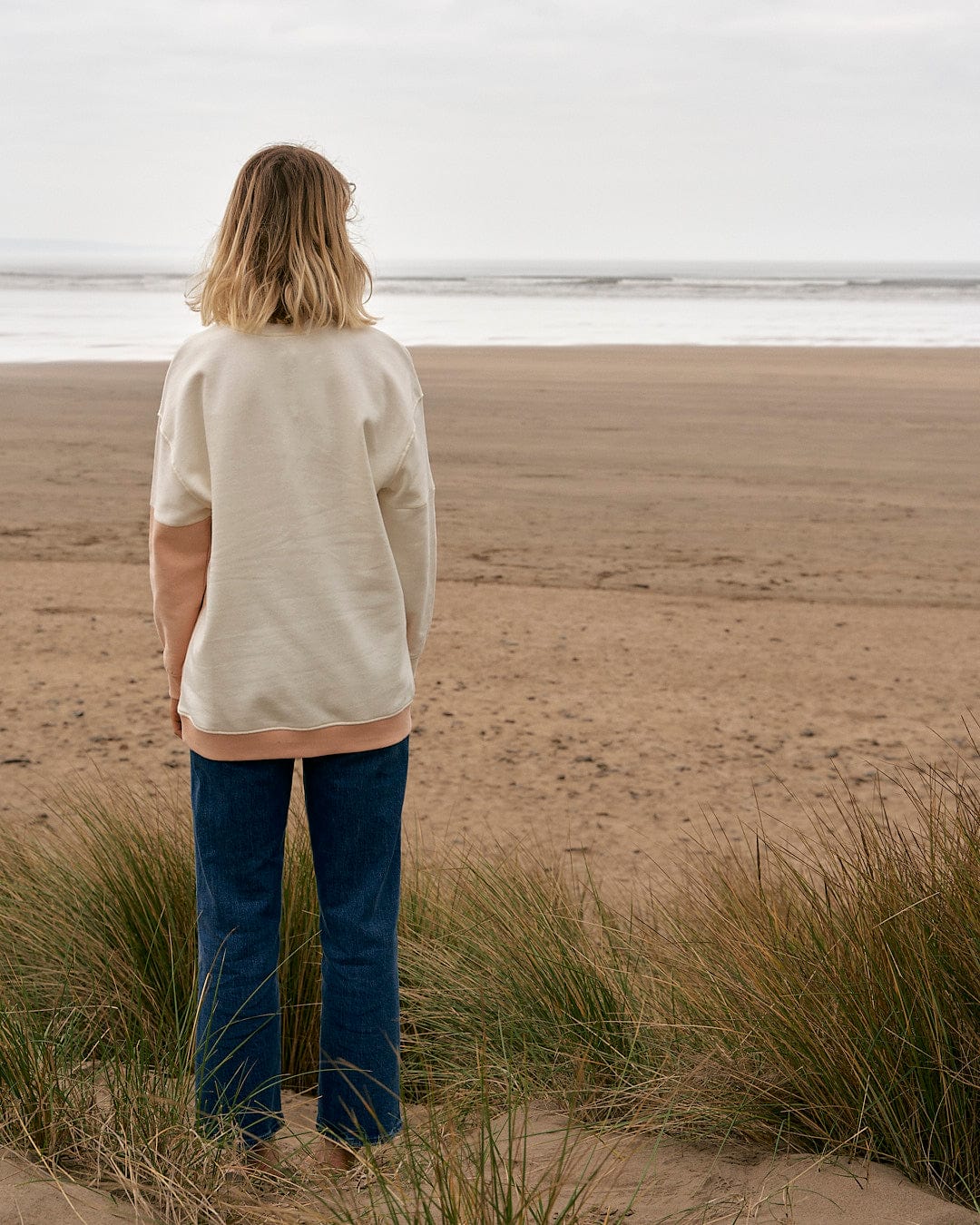 A woman standing on a beach looking at the ocean, wearing the Sofie - Womens Boyfriend Fit Sweat in Light Orange/Cream by Saltrock.