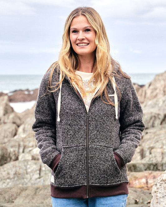 A woman wearing a Saltrock Sofie - Womens Borg Lined Zip Hoodie - Brown standing on rocks near the ocean.