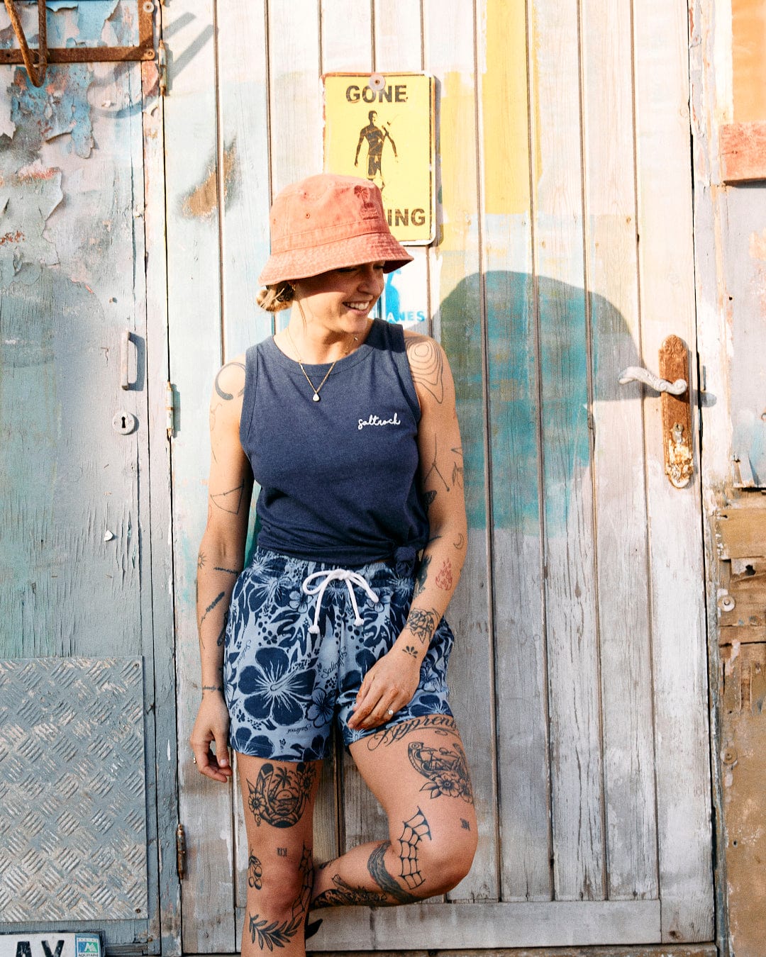 A person with tattoos, wearing a tank top and Saltrock Soifra Hibiscus - Womens Sweat Short - Blue with an elasticated waist and a peached soft hand feel finish, stands smiling in front of a rustic wooden door with a "Gone Surfing" sign.