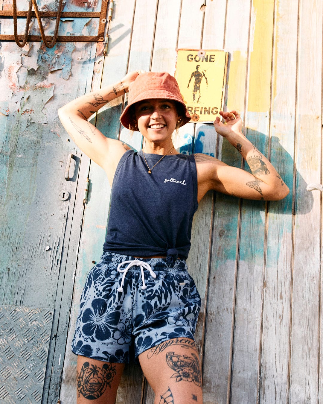 A person wearing a red bucket hat, blue tank top, and Soifra Hibiscus - Womens Sweat Short - Blue by Saltrock with an elasticated waist, posing in front of a weathered wall with a "Gone Surfing" sign. The individual has multiple tattoos on their arms and legs, adding a touch of Hawaiian hibiscus flair to their laid-back style.