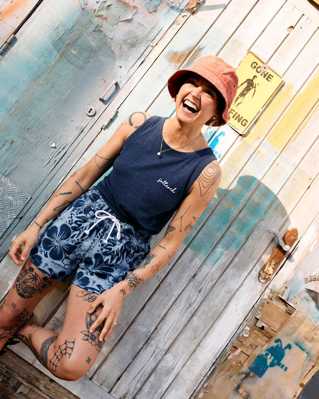 Person wearing a bucket hat, tank top, and Soifra Hibiscus - Womens Sweat Short - Blue from Saltrock with an elasticated waist, smiling against a weathered wooden wall with tattoos visible and a "Gone Surfing" sign in the background. The Hawaiian hibiscus design adds to the tropical vibe.