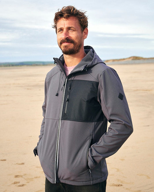 Description (modified): A man standing on a beach wearing a dark grey Saltrock Munros - Mens Softshell Jacket made from water-resistant fabric, with zip pockets.