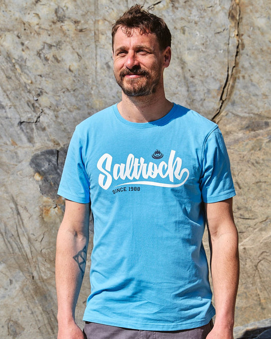 A man wearing a Speed - Mens Short Sleeve T-Shirt - Blue by Saltrock standing in front of a rock.