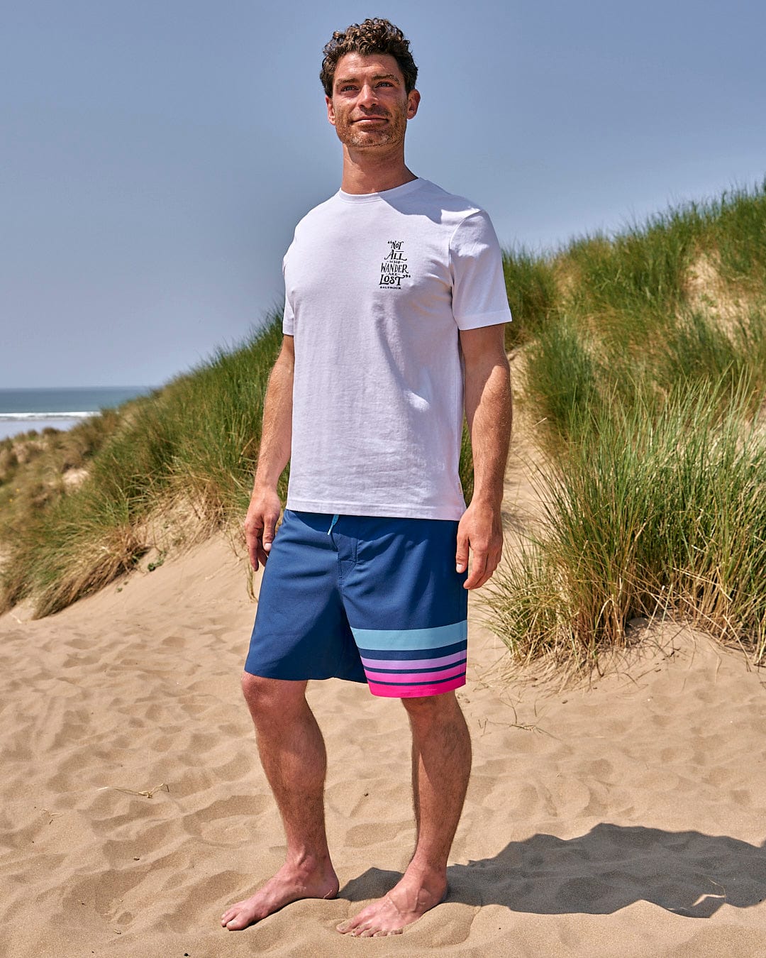 A man standing on a sand dune wearing Saltrock Splash Gradient - Mens Boardshort - Dark Blue t-shirt and shorts.