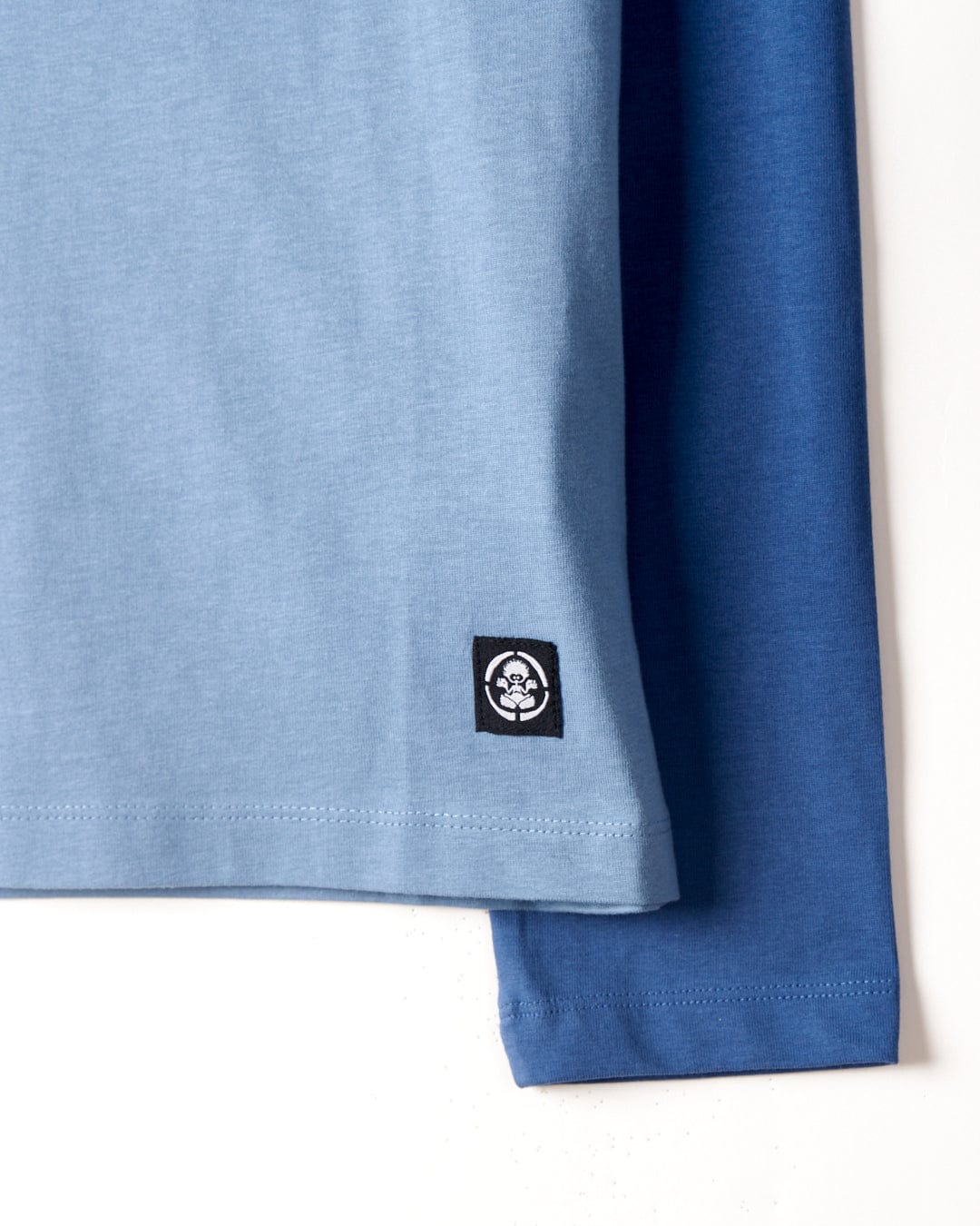 Close-up of a blue Spray Stripe - Kids Raglan Long Sleeve T-shirt with raglan contrast sleeves and a small black Saltrock branding patch on the left side.