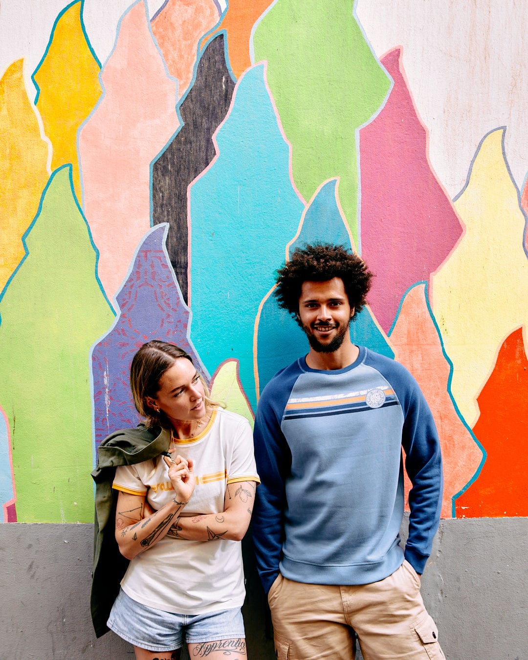 A man and a woman leaning against a colorful graffiti wall, the woman looking at the man who is smiling at the camera, both wearing Saltrock Spray Stripe - Recycled Mens Sweat - Blue shirts with a crew neckline.