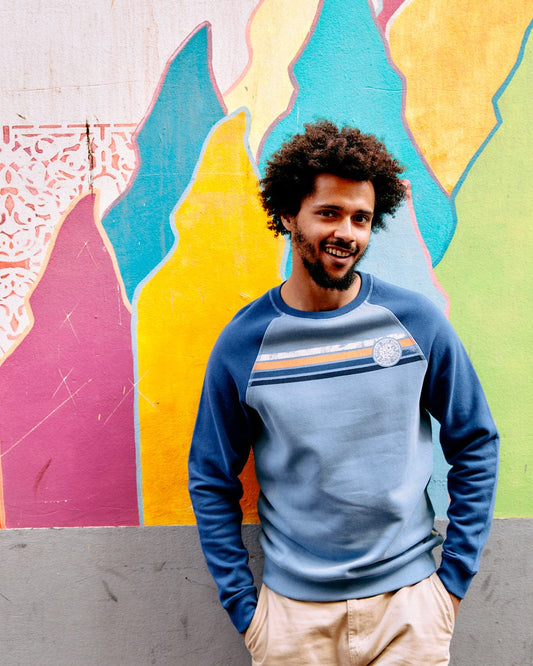 A man with an afro smiling, wearing a Saltrock blue Spray Stripe - Recycled Mens Sweat with a crew neckline, standing in front of a colorful graffiti wall.