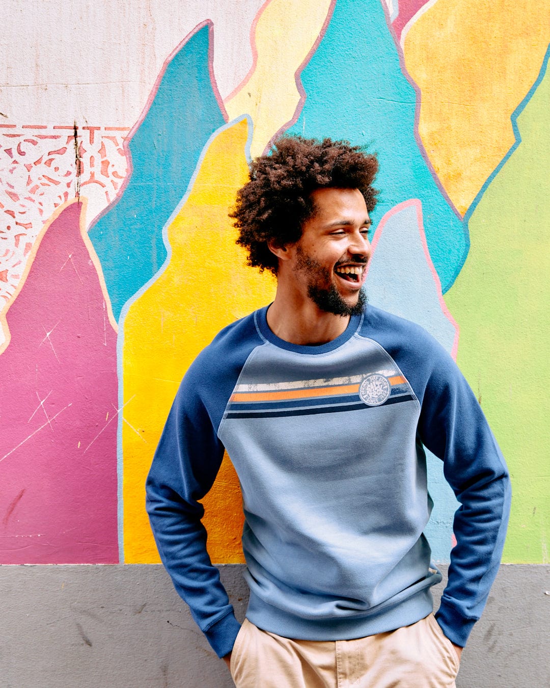 A man with an afro smiling, standing in front of a colorful graffiti wall. He is wearing a Saltrock Spray Stripe - Recycled Mens Sweat in Blue with raglan sleeves and beige pants.