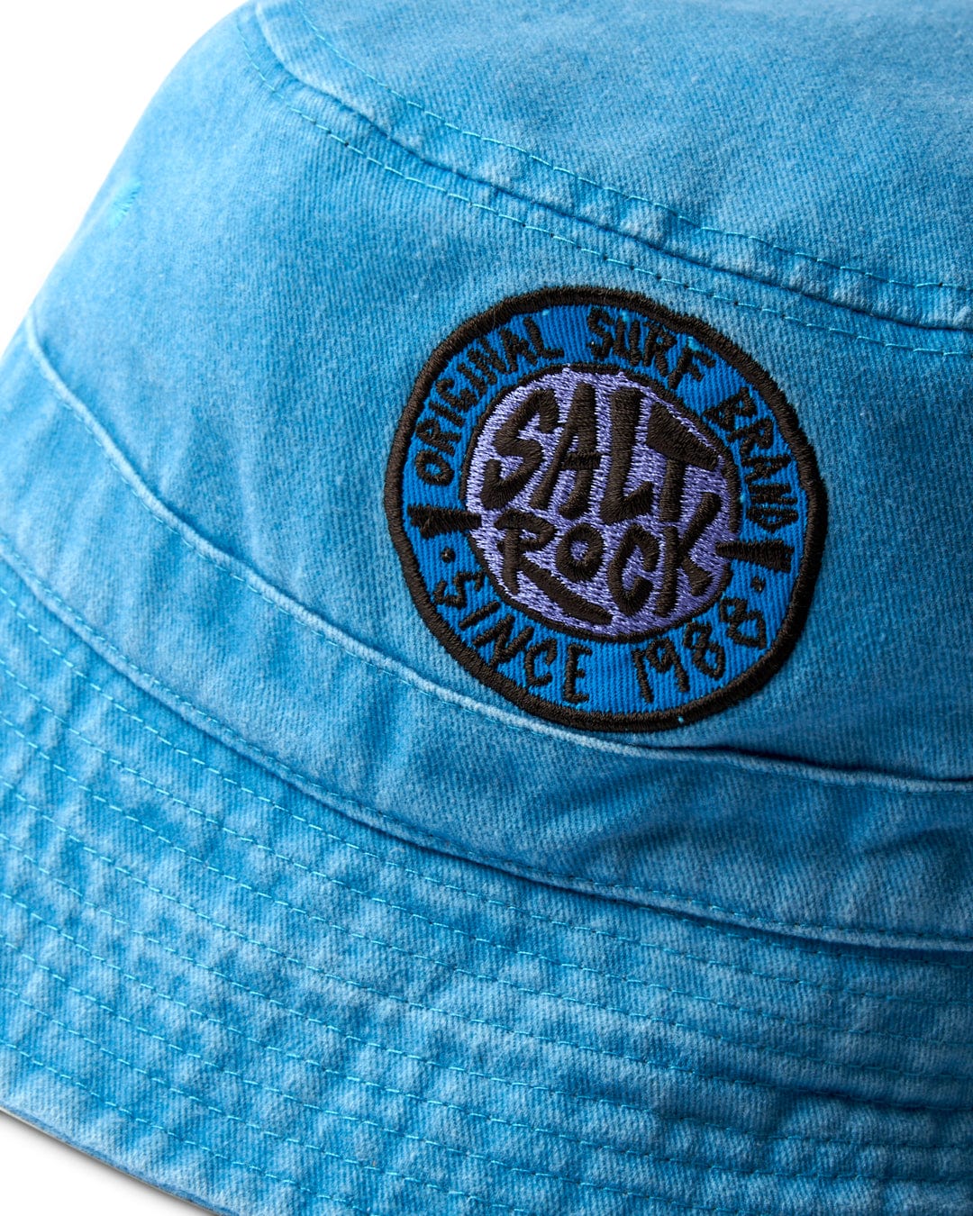 Close-up of a blue 100% cotton fabric cap with an embroidered Saltrock badge that reads "Original Surf Since 1983" on the SR Original - Bucket Hat - Blue.