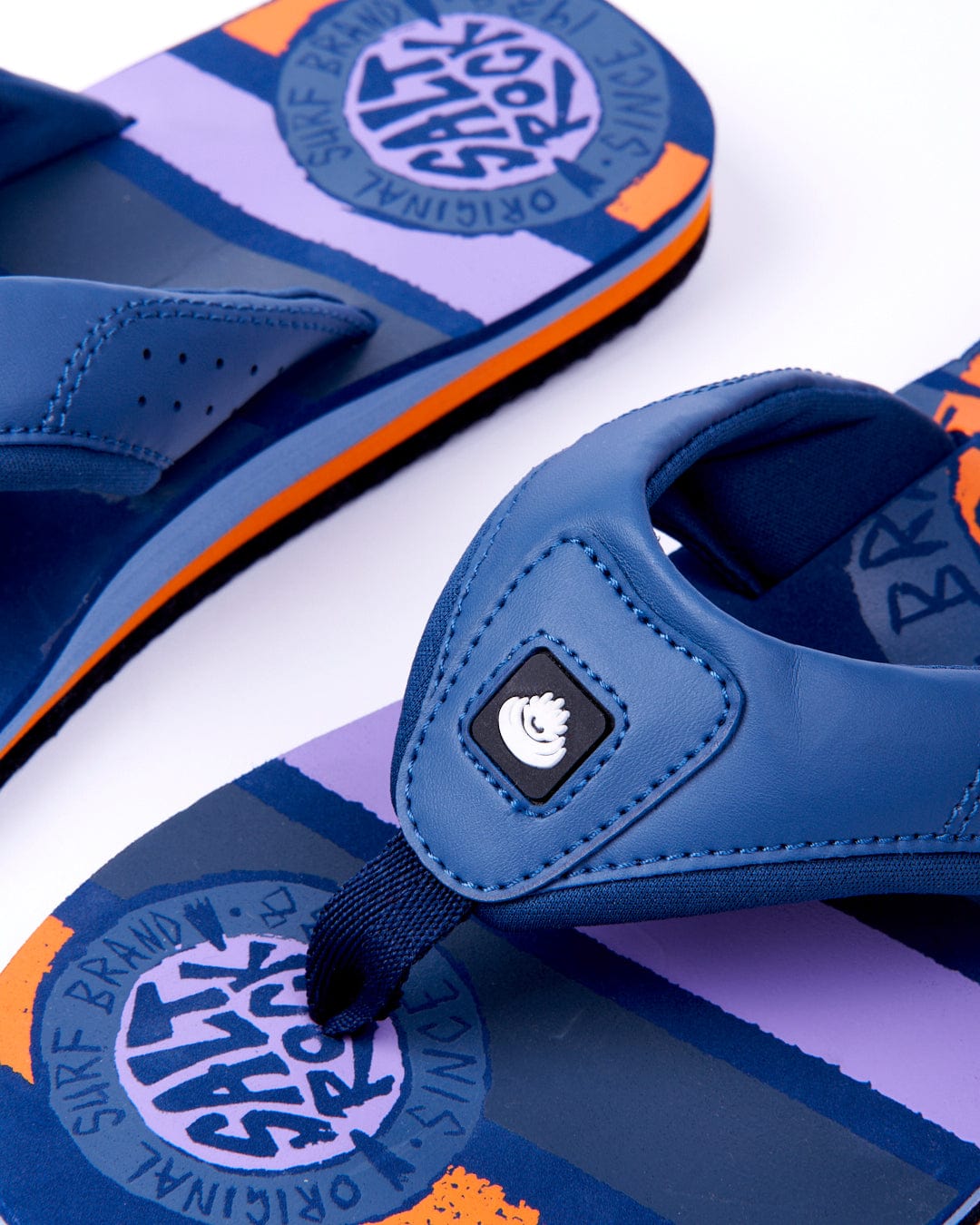 A close-up of blue flip-flops with orange accents and Saltrock branding on the insoles.