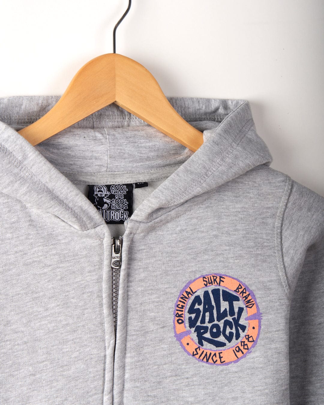 Close-up of a Saltrock SR Original - Kids Zip Hoodie - Grey with a "Salt Rock Original Surf Brand Since 1988" logo on the chest, hanging on a wooden hanger.