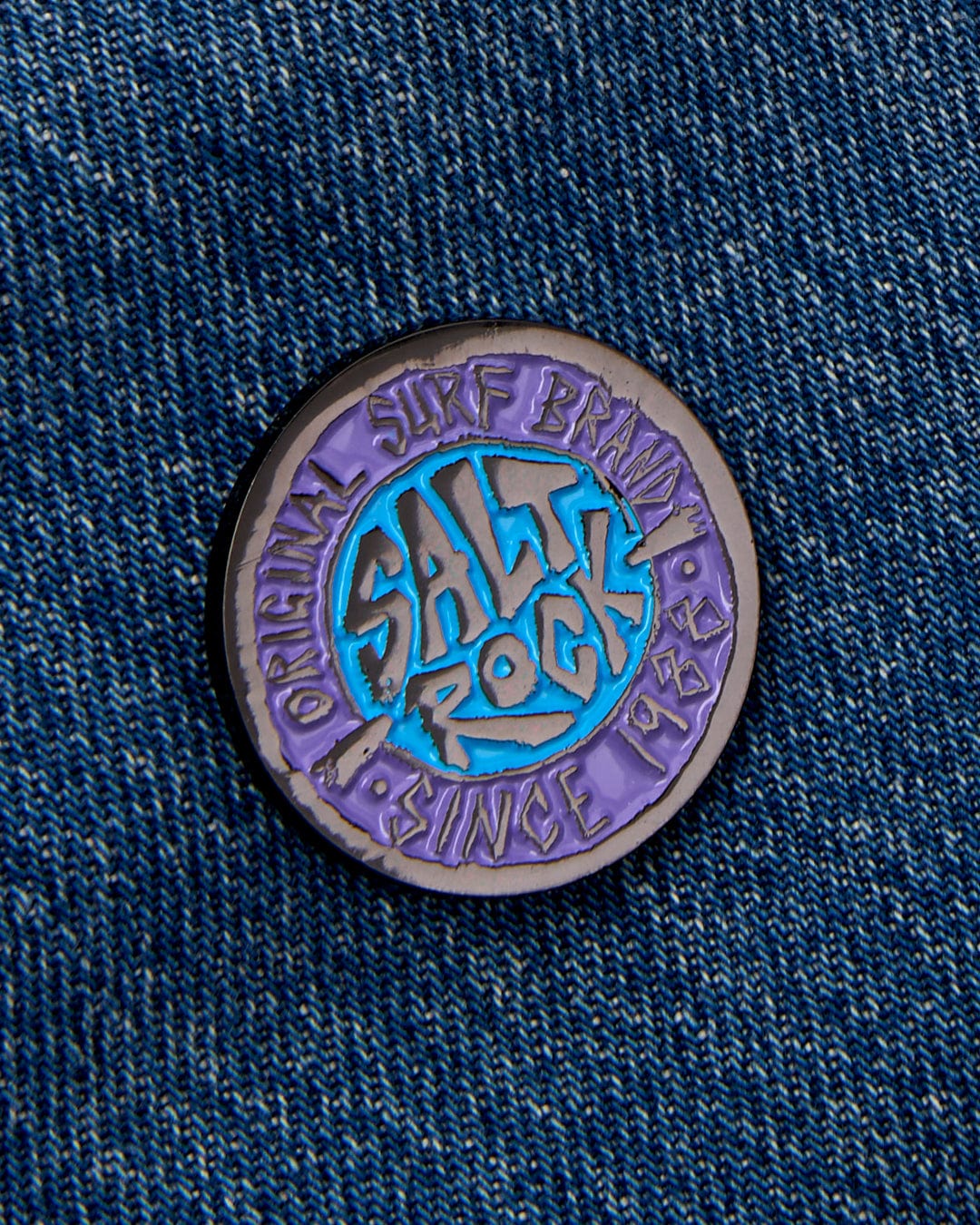 A SR Original pin badge featuring Saltrock graphics, pinned on a denim fabric with a safety clasp.