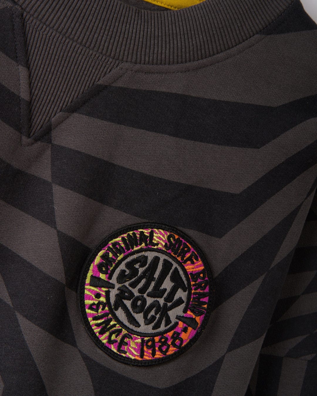 Close-up of the SR Warp - Men's Oversized Sweat in grey by Saltrock, showcasing black stripes and a round patch reading "Salt Rock" and "ORIGINAL SURF BRAND SINCE 1988.