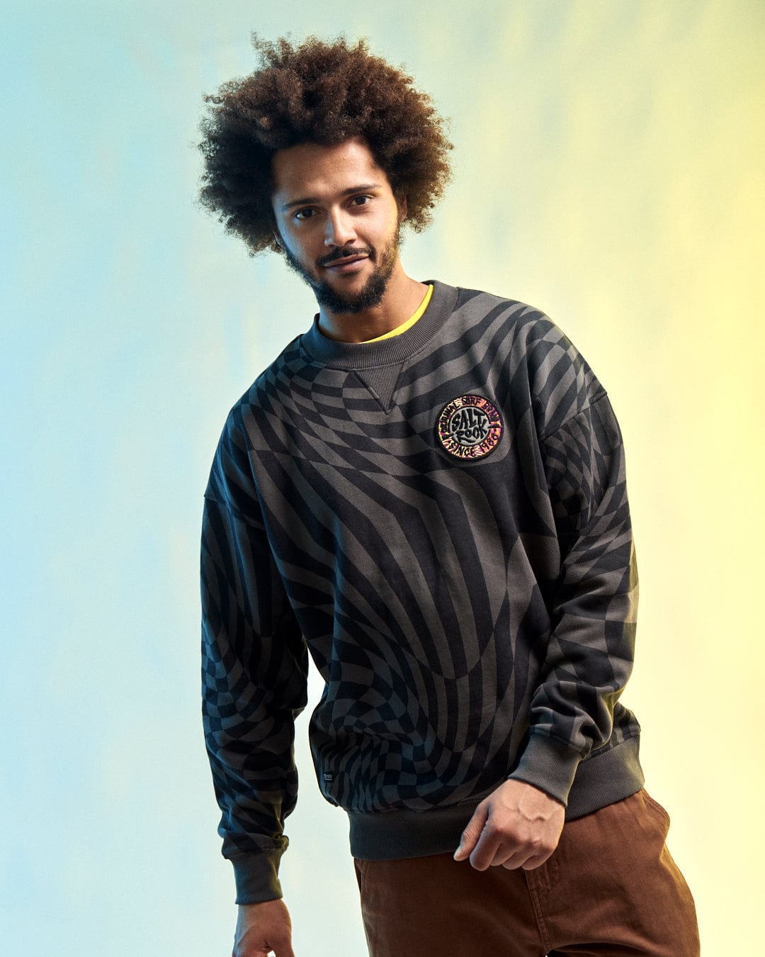 A man with curly hair stands against a blue and yellow gradient background, wearing Saltrock's SR Warp - Men's Oversized Sweat in grey.