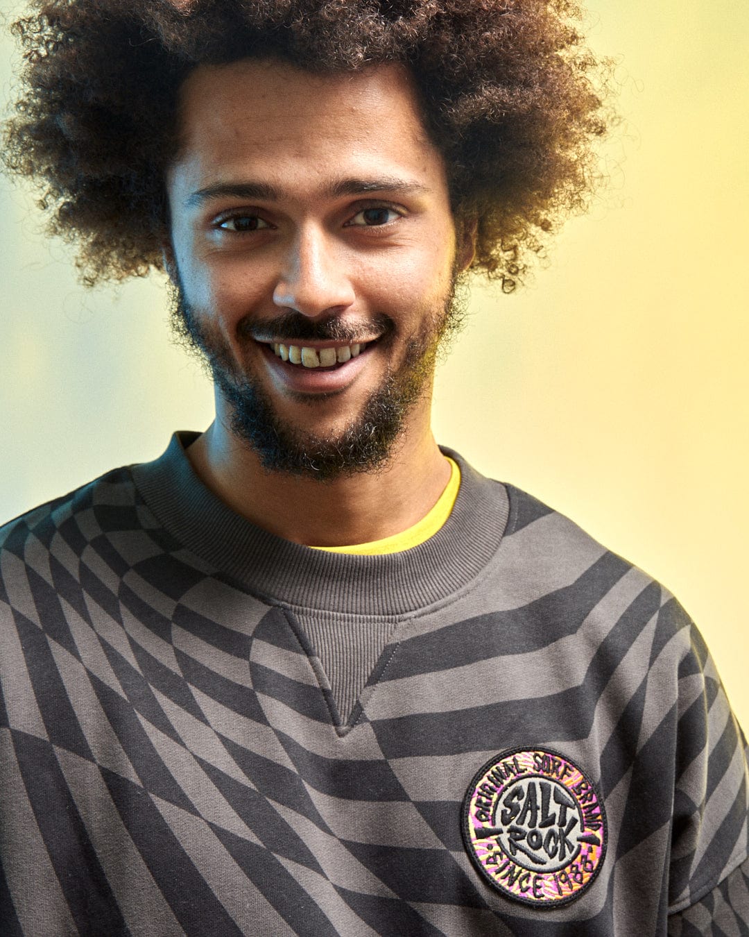 A person smiles in Saltrock's SR Warp Men's Oversized Sweat in grey featuring a geometric print and "CALL ROCK" patch, set against a yellow background with soft lighting.