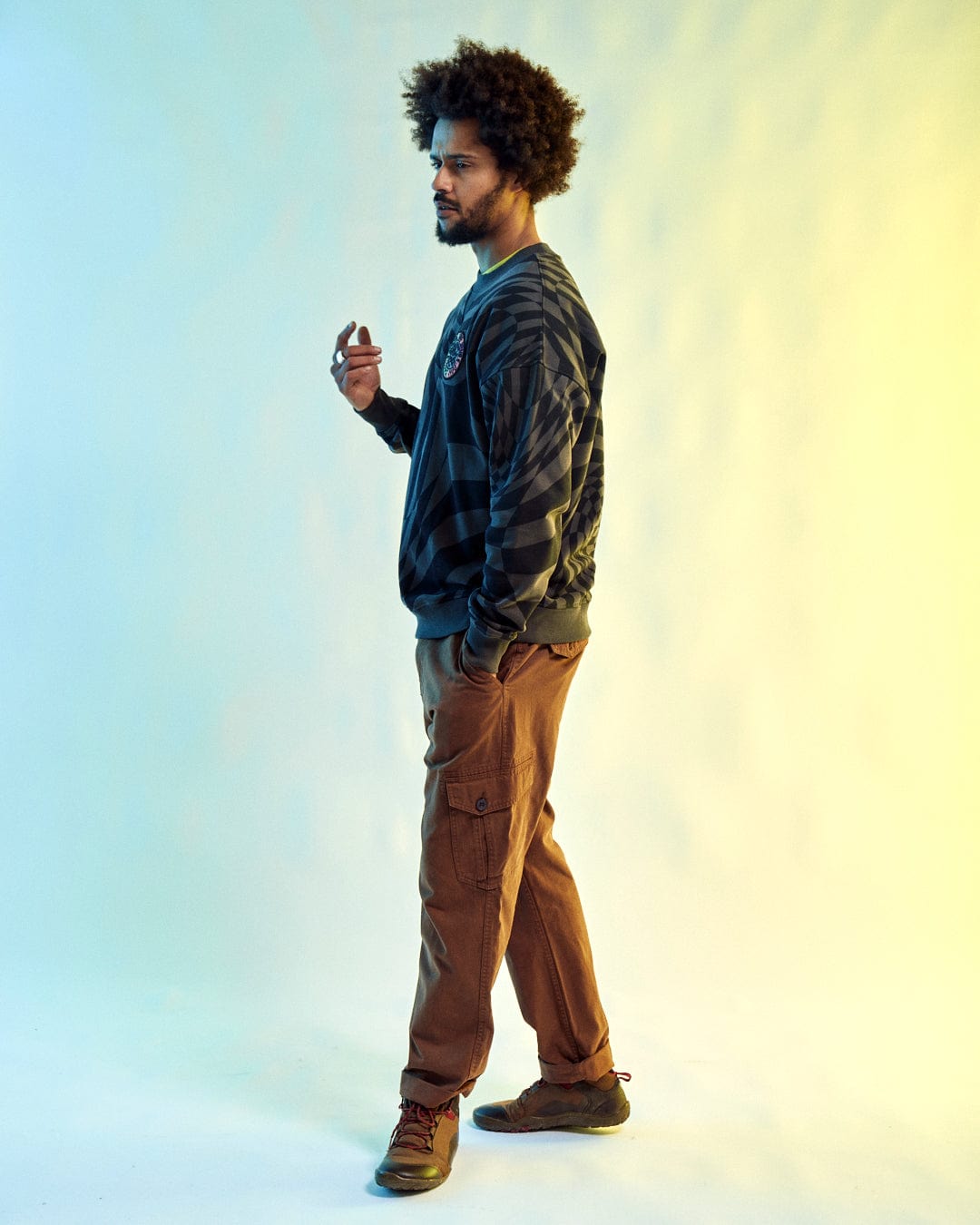 A man with an afro sports the SR Warp - Mens Oversized Sweat in grey by Saltrock and brown cargo pants, set against a gradient backdrop. His outfit is stylishly complemented as he holds a small recycled item.