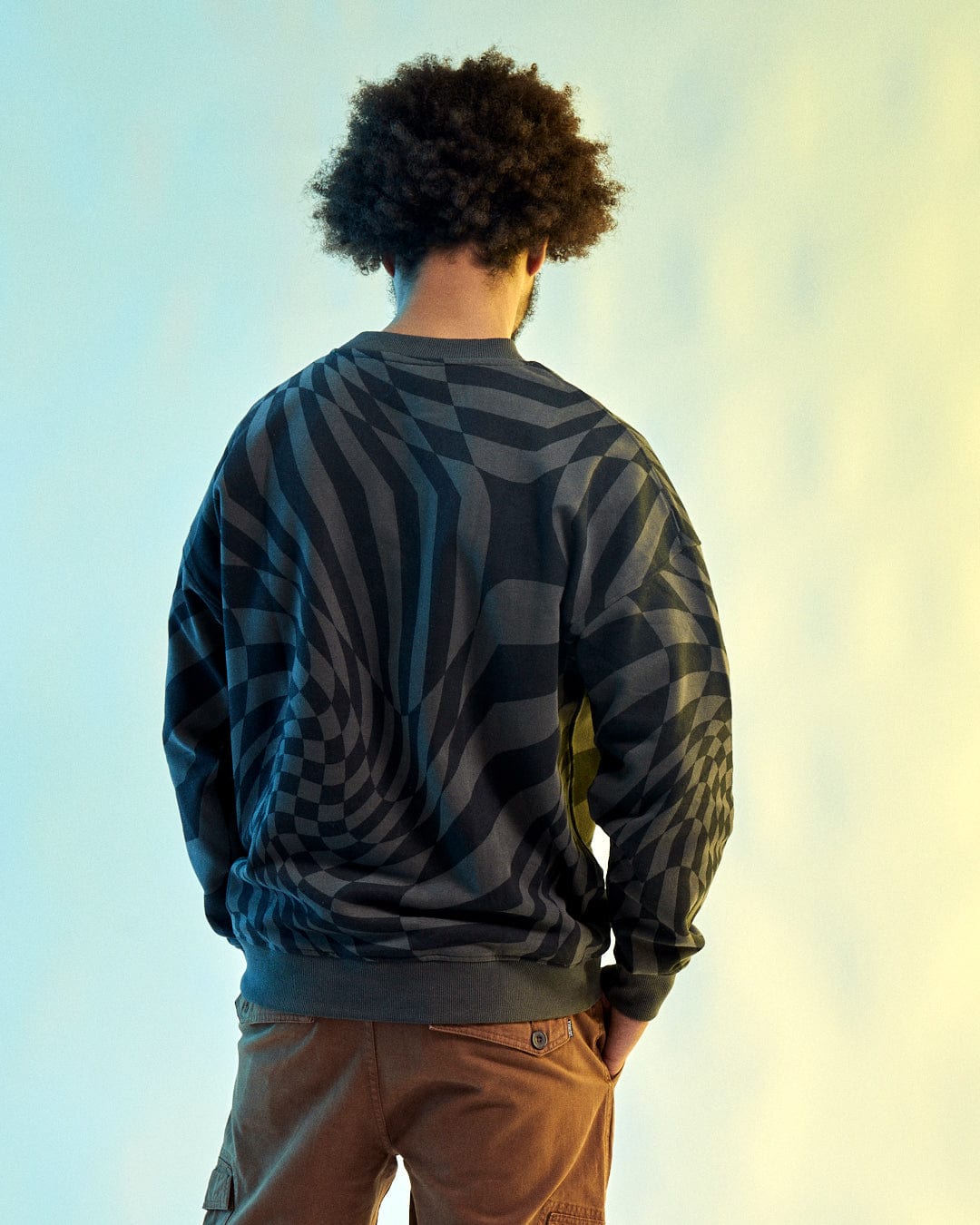 A person with curly hair facing away wears the SR Warp - Mens Oversized Sweat in grey by Saltrock, featuring a black and gray geometric pattern, set against a gradient background.
