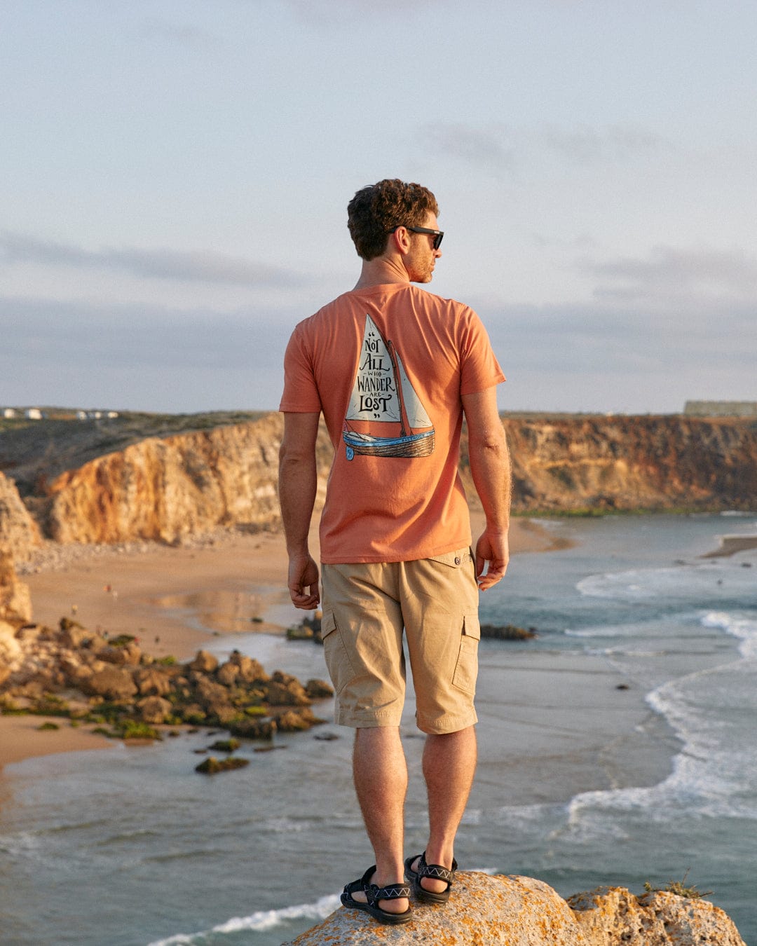 A man standing on top of a cliff, gazing out at the vast ocean wearing a Saltrock Lost Ships - Mens Short Sleeve T-Shirt in Orange.