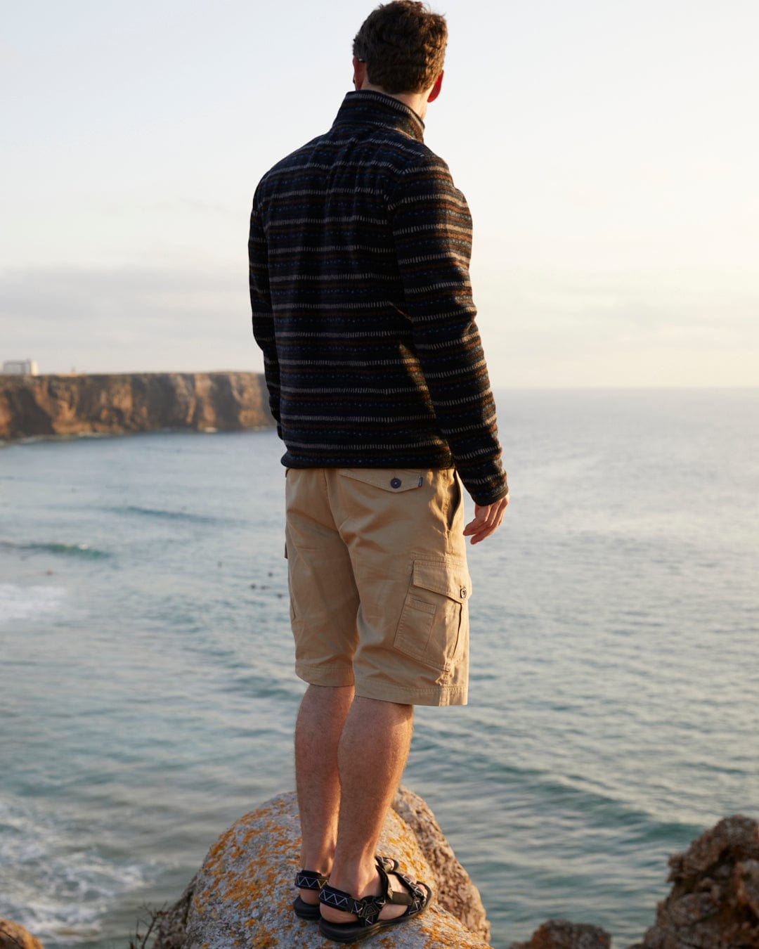 Man standing on a rock overlooking the ocean at sunset, his Saltrock Penwith II - Mens Cargo Shorts in Cream subtly outlined against the fading light.