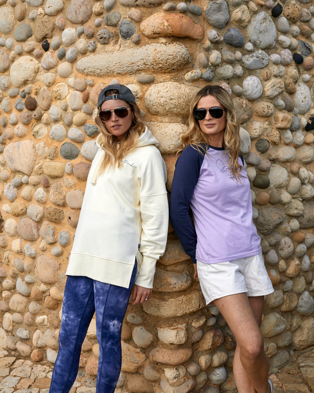 Two women posing in casual Saltrock branded attire against a stone wall featuring the Poster - Womens Pop Hoodie - Cream.