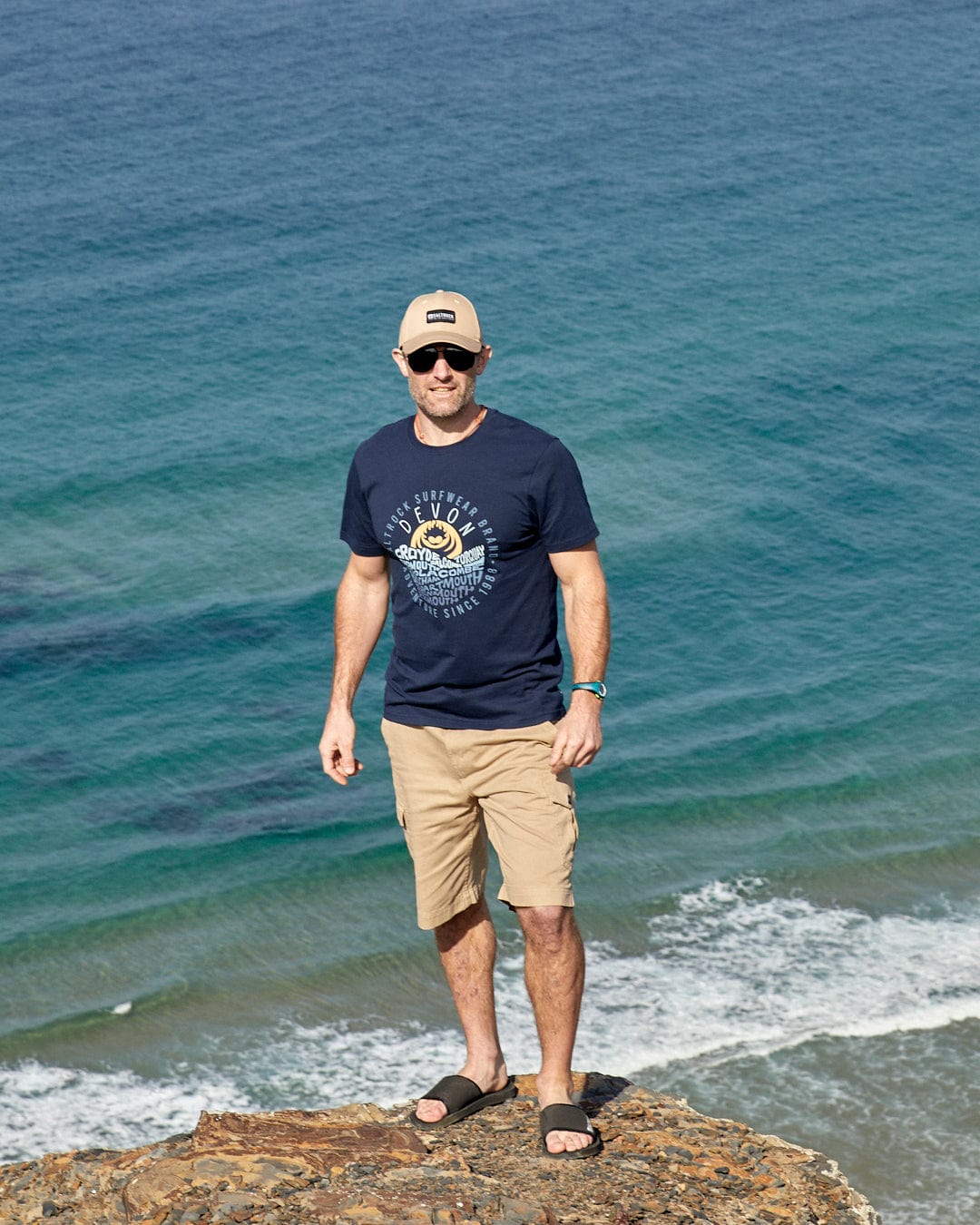 A man standing on a cliff wearing Saltrock Layers Devon - Mens Short Sleeve T-Shirt in Blue, overlooking the ocean.