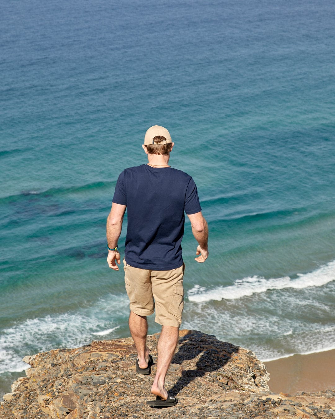 A man, wearing Layers Devon - Mens Short Sleeve T-Shirt in Blue from Saltrock, is standing on a cliff overlooking the ocean.
