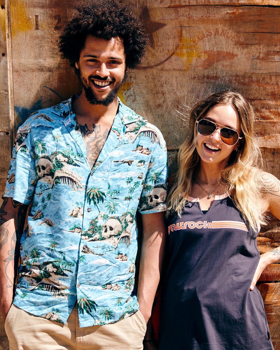 A man and a woman smiling in casual summer clothes, featuring Hawaiian Isle - Mens Short Sleeve Shirt in Blue by Saltrock, standing in front of a rustic wooden background.