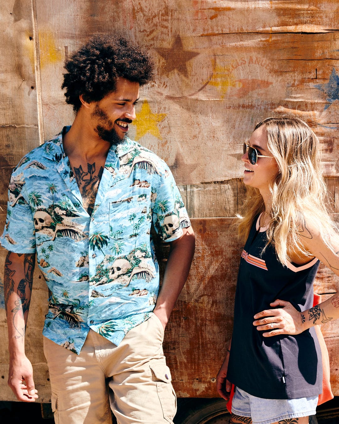 A man and a woman smiling at each other, standing by a textured wooden wall. The man wears a Saltrock Hawaiian Isle - Mens Short Sleeve Shirt in Blue; the woman sports a black tank top.