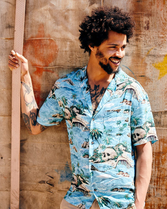 A man with an afro and tattoos smiles while leaning against a graffiti-covered wall, wearing a Saltrock Hawaiian Isle Men's Short Sleeve Shirt in Blue.