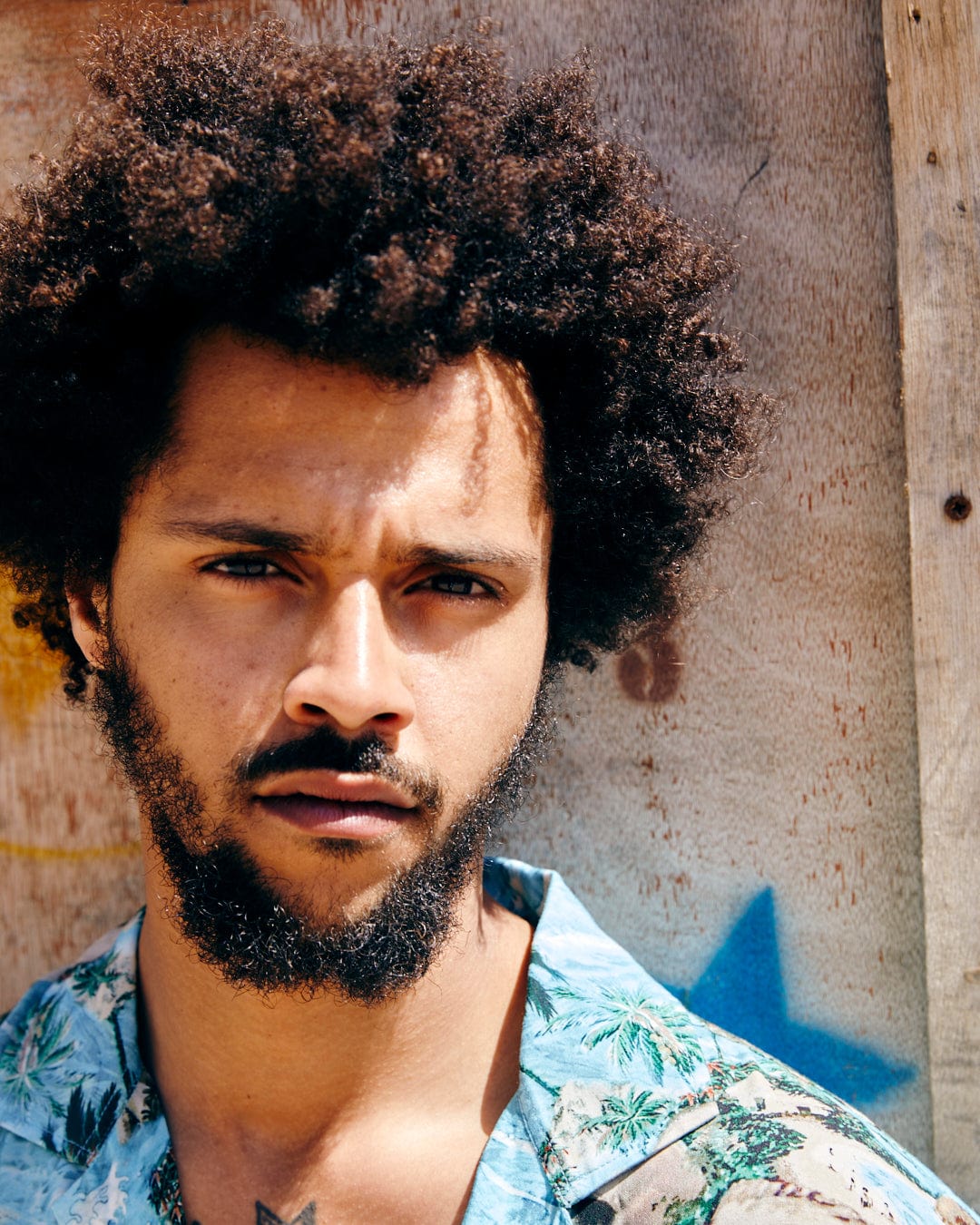 Close-up portrait of a young man with a curly afro and beard, wearing a Saltrock Hawaiian Isle Men's Short Sleeve Shirt in Blue, standing against a wooden backdrop.
