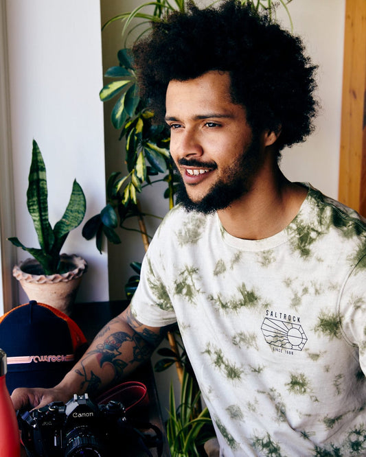 A young man with a curly afro smiles gently, sitting by a window with plants and a camera, wearing the Saltrock Vantage Outline - Mens Tie Dye T-Shirt in Green.