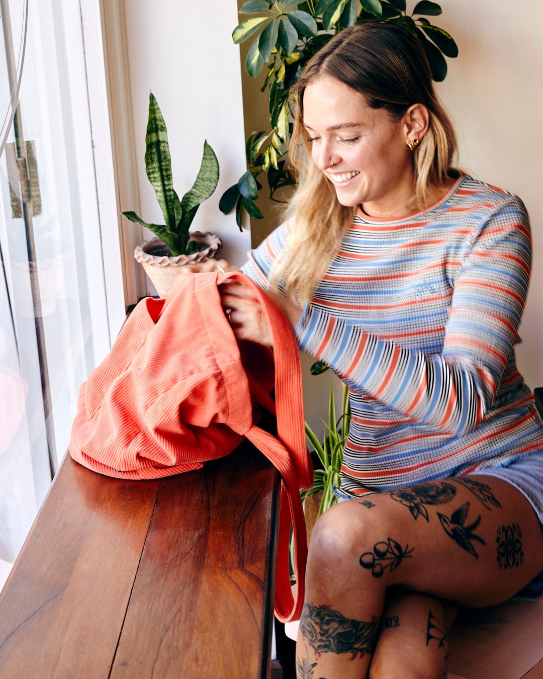 A woman with tattoos, smiling as she packs a Lanie - Womens Long Sleeve T-Shirt in Red into a red backpack at a wooden table near a window, the bag prominently featuring Saltrock branding.