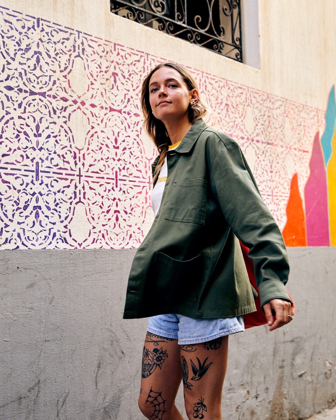 A woman in a Saltrock Barden - Womens Lightweight Utility Jacket in Dark Green and denim shorts stands confidently in front of a colorful mural on an urban street.