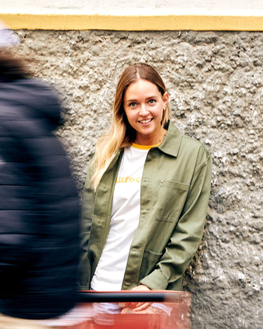 A smiling woman in a Saltrock Barden - Womens Lightweight Utility Jacket - Dark Green and white shirt seated by a grey wall, with a blurred figure passing by.