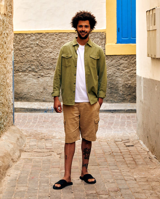 A young man with curly hair stands in a narrow alley, wearing a Penare - Mens Long Sleeve Shirt in Green by Saltrock, tan shorts, and black sandals, smiling at the camera.