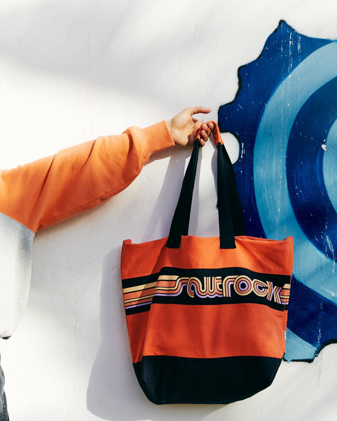 A person in an orange jacket with durable hand covers reaching for a Saltrock Cora Retro Beach Bag - Coral hanging on a white wall with blue graffiti.