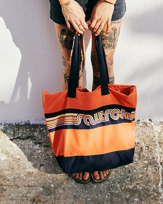 A person holding a Cora Retro Beach Bag in Coral from Saltrock, with durable material and the word "superfolk" on it, standing against a white wall, showcasing tattooed legs and feet in sandals.