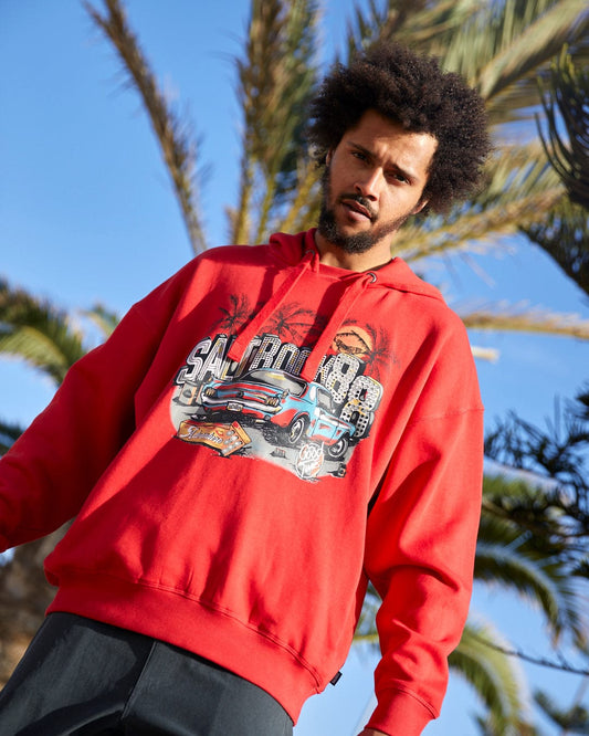 A man in a Neon Boneyard - Mens Pop Hoodie in Red, by Saltrock, stands under palm trees, looking at the camera.