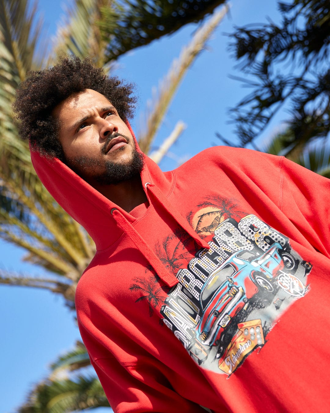 Man in Neon Boneyard - Mens Oversized Pop Hoodie - Red by Saltrock, with a graphic design, looking away, standing under blue sky with palm trees in the background.