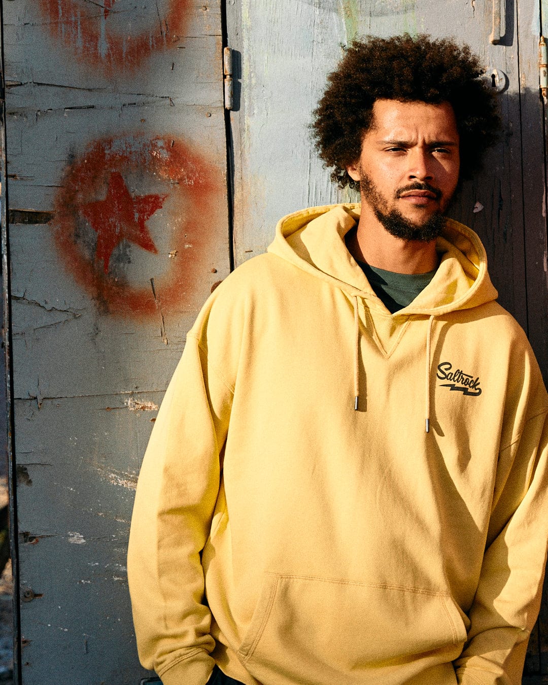 Man with curly hair standing by a weathered door with a red star graffiti, wearing a yellow Saltrock Gas Station - Recycled Mens Pop Hoodie.