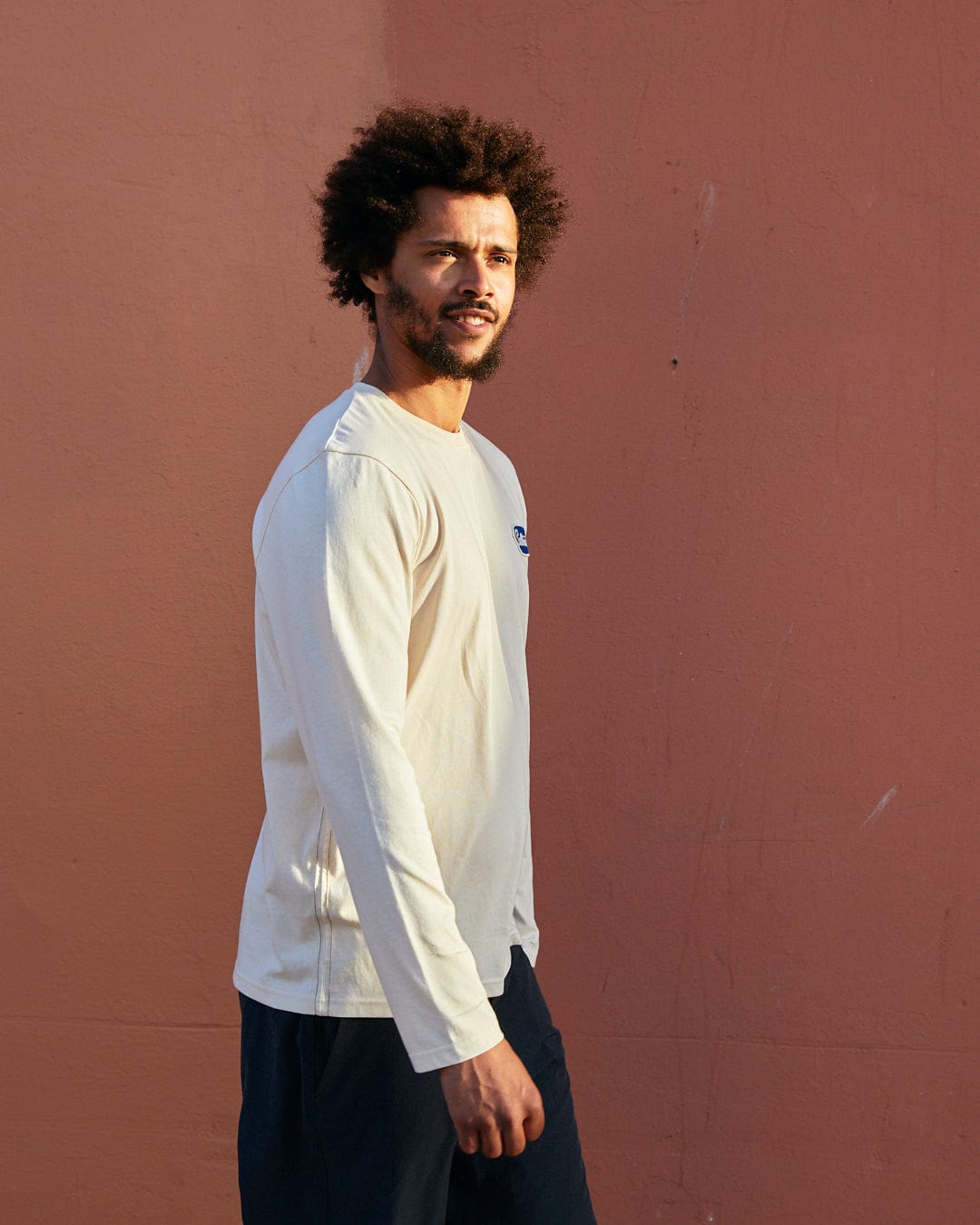 A man with an afro standing sideways in front of a pink wall, wearing a cream Saltrock long-sleeve shirt and navy shorts, detailed with a nylon Saltrock badge.