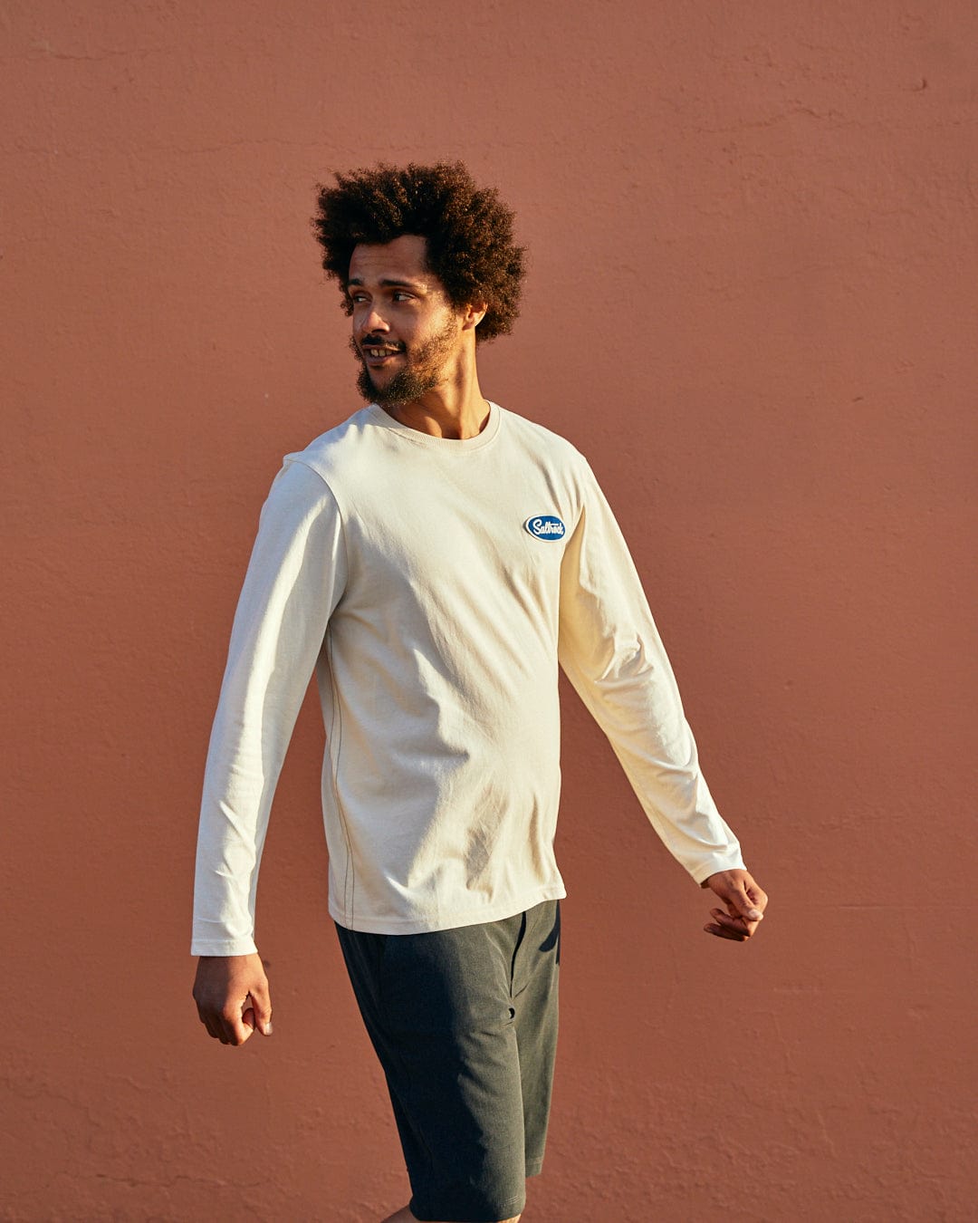 A man with an afro walking past a terracotta wall, wearing a white long-sleeve shirt with a blue Nylon Saltrock badge and dark pants, looking to the side wearing the Saltrock Attendant Mens Long Sleeve T-Shirt in Cream.