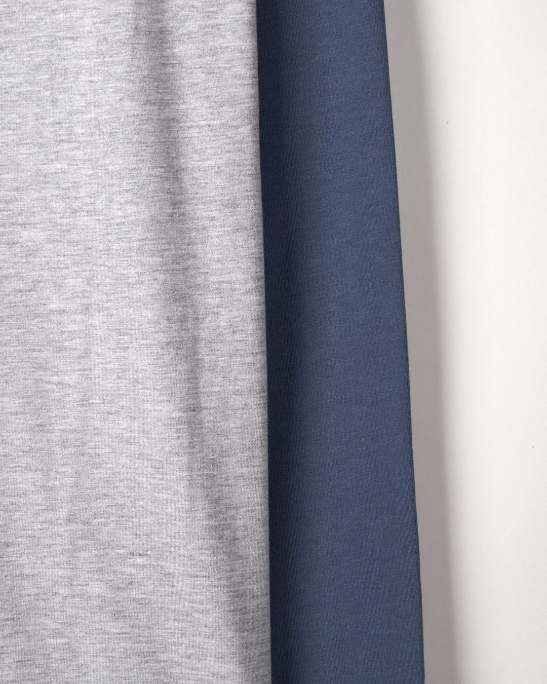 Close-up of two fabrics, one Strike Logo - Mens Long Sleeve Raglan T-Shirt - Grey Marl from Saltrock and textured, the other dark blue and smooth, laid side by side on a flat surface.
