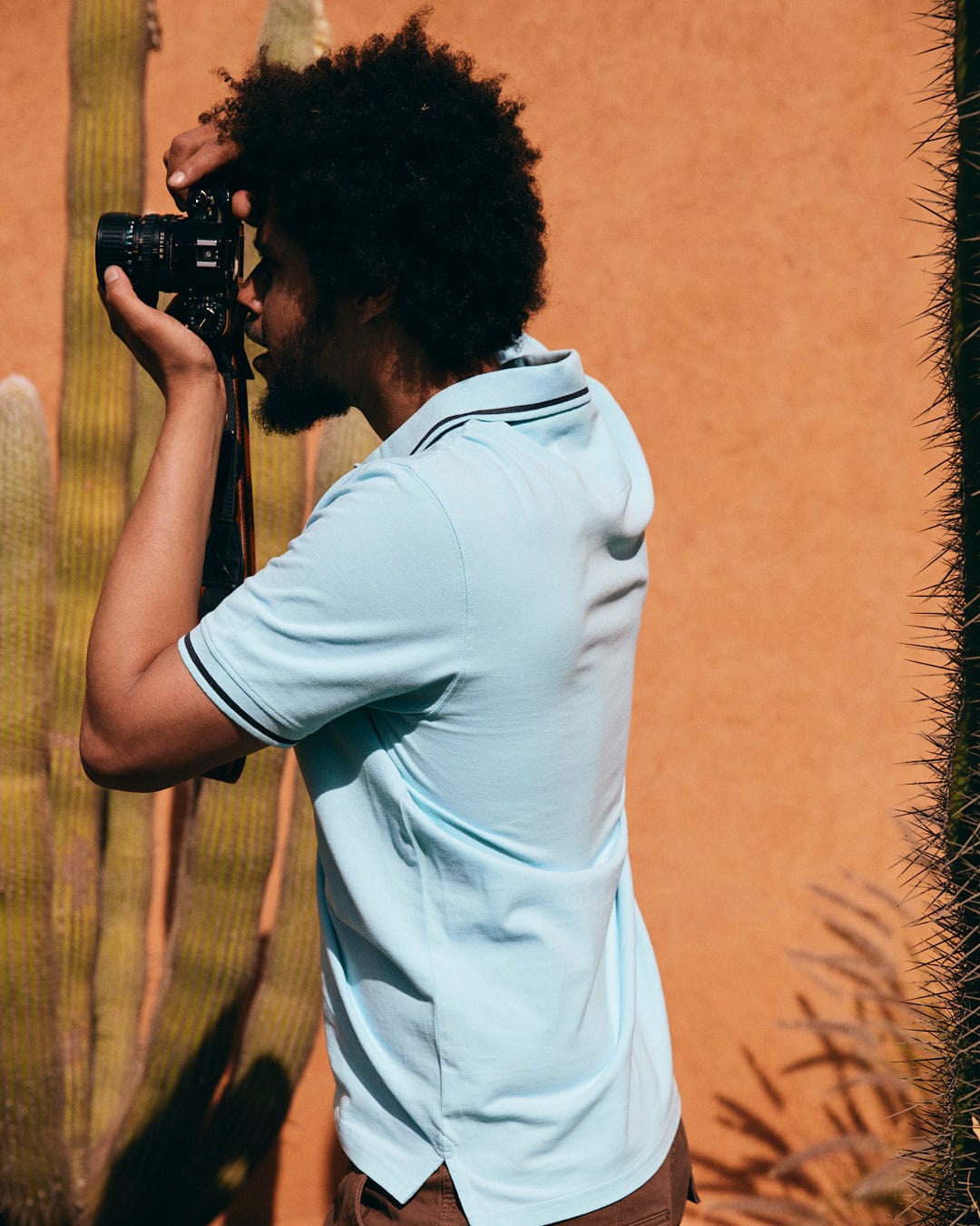 A person wearing the Strike Logo - Mens Short Sleeve Polo Shirt - Light Blue by Saltrock and brown pants is holding a camera up to their eye, capturing a photo next to a cactus.