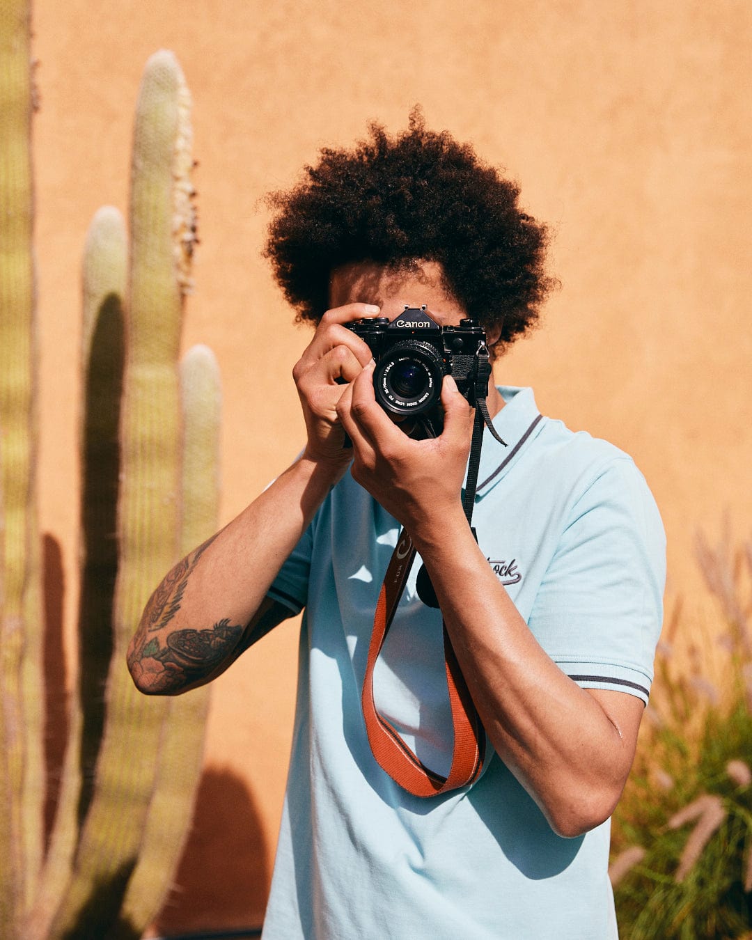 Person in a Strike Logo - Mens Short Sleeve Polo Shirt - Light Blue by Saltrock taking a photo with a camera in front of a tall cactus and an orange wall.